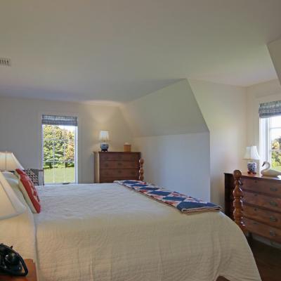 Cozy bedroom with a quilted bed, wooden dressers, and bright windows overlooking a garden.