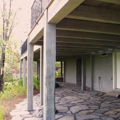 Under a modern concrete structure, a stone pathway leads to a rustic, wooded area.