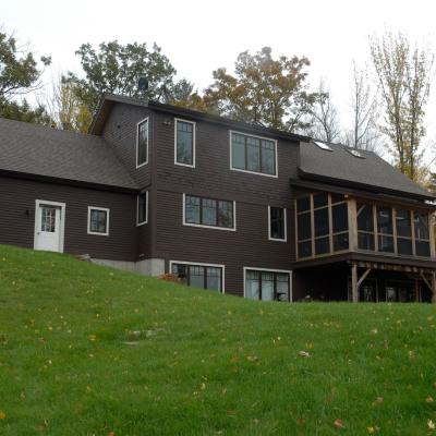 A modern brown wooden house with a screened porch and large windows, set on a grassy slope.