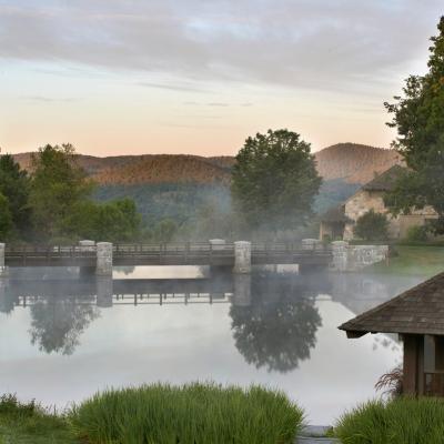 Fog envelops a tranquil pond with a bridge, surrounded by lush greenery and mountains.