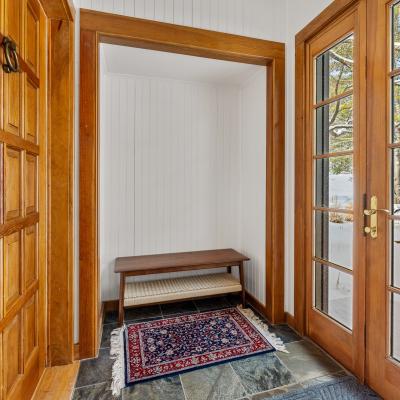 Bright entryway with wooden doors, a bench, and a rug, leading to a snowy outdoor view.