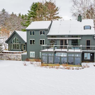 Gray house with snowy surroundings and a large deck, surrounded by trees.