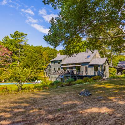 Spacious house with a deck, pool, and surrounded by lush trees under a clear blue sky.