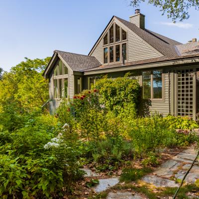Cozy gray home surrounded by lush greenery and colorful flowers under a clear blue sky.