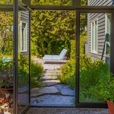 View through a glass door to a garden path and lounge chairs surrounded by greenery.