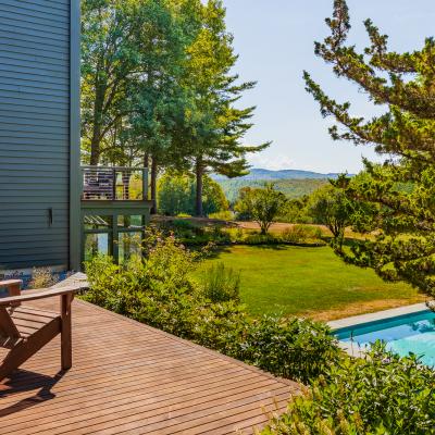 Wooden deck with chairs overlooking a lush garden and pool against a mountain backdrop.