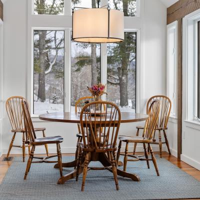 Cozy dining area with a round table, wooden chairs, and large windows showing a snowy landscape.