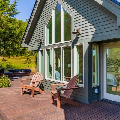 Modern cabin exterior with a deck, two chairs, and a hot tub surrounded by greenery.