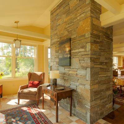 Cozy living space with stone fireplace, armchair, and large windows overlooking greenery.