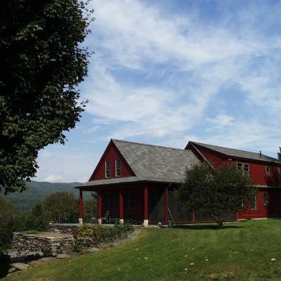 Red farmhouse with a porch, surrounded by trees and mountains under a blue sky.