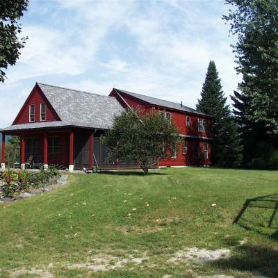 Red house with a porch, surrounded by trees and a grassy lawn under a blue sky.