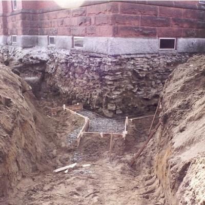Excavated area beside a stone building, showing foundation work and soil.