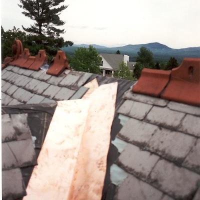 View of a slate roof with copper flashing and a cloudy mountain backdrop.