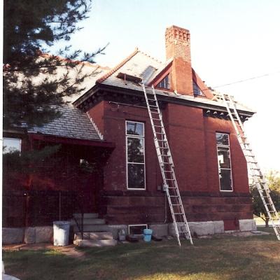 Red brick house with ladders leaning against it, under renovation, surrounded by grass.